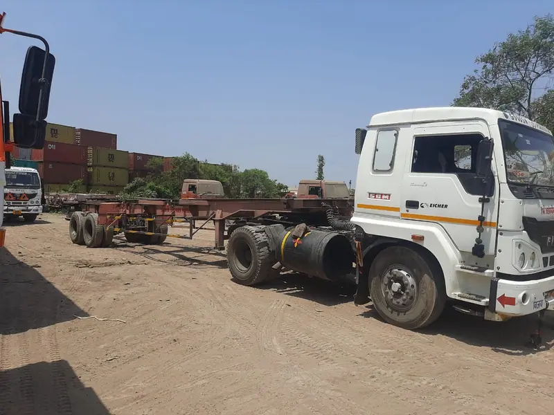 Tractor with an empty skeletal trailer in a yard, with stacked containers in the background