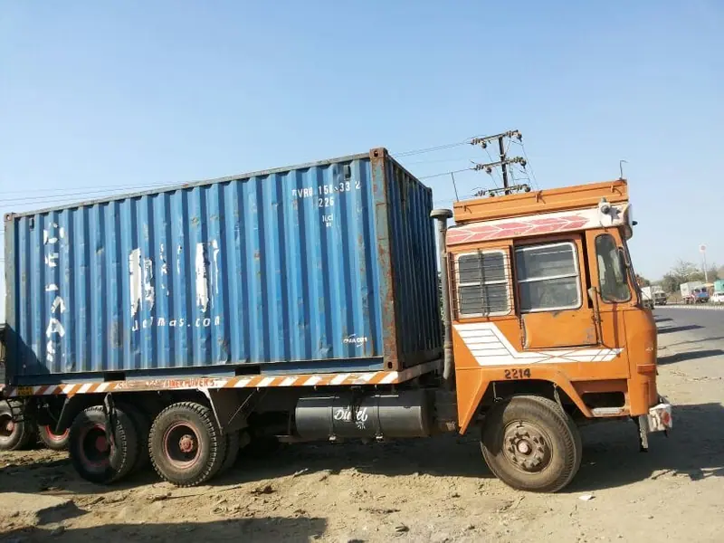 Orange truck carrying a blue 20ft container across a dusty yard